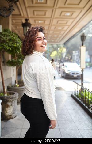 Seitenansicht der prallen Frau trägt trendige Kleidung im Stehen Stadt am sonnigen Tag und selbstbewusst Blick auf die Kamera Stockfoto