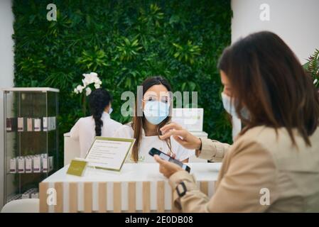 Weibliche Rezeptionistin in Maske, die an der Theke im modernen Schönheitssalon steht und auf die Zahlung von Frau für die Hautpflege wartet Stockfoto