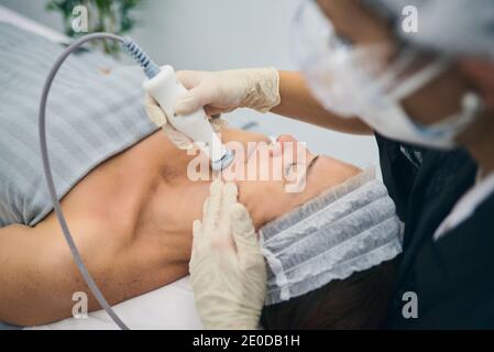 Professioneller Dermatologe, der während der Hautpflegebehandlung in einer Schönheitsklinik ein HF-Hebegerät auf das Gesicht einer reifen Frau anwendet Stockfoto