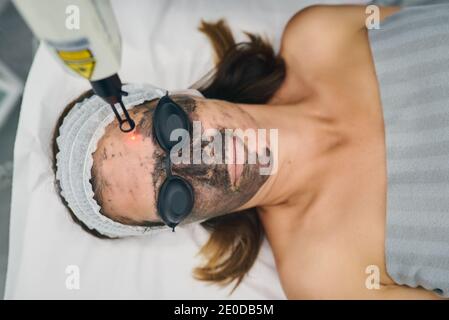 Von oben anonymen Kosmetikerin, die Maschine für Carbon Peel On verwendet Gesicht der weiblichen Klientin, die in einer Schutzbrille in Schönheit liegt Klinik Stockfoto