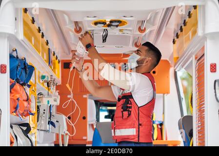 Seitenansicht des schweren männlichen Sanitäters in medizinischer Maske stehend Mit Dropper im modernen Ambulanzwagen Stockfoto