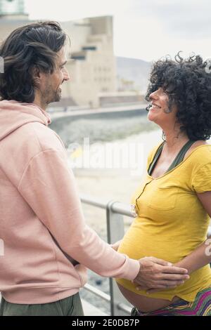 Lächelndes Paar wartet auf ein neues Familienmitglied am Meer an einem windigen Tag auf Gran Canaria, Spanien. Glücklicher Vater hält den Bauch der Mutter Stockfoto