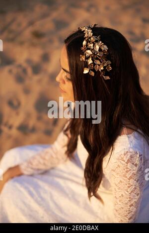 Von oben Seitenansicht der jungen romantischen Bohemian Brünette in Weißes Hochzeitskleid und goldener Blumenkranz auf Sand sitzend Stockfoto