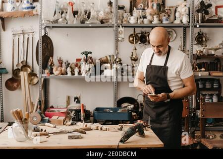 Serious mittleren Alters männlichen Zimmermann in Schürze Messaging auf Handy Telefonieren Sie in der Nähe der Werkbank mit professionellen Tools in kreativ Arbeitsstudio Stockfoto