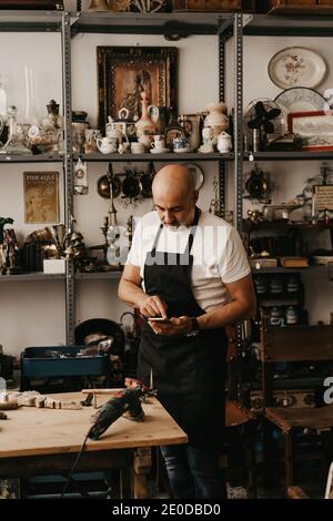 Serious mittleren Alters männlichen Zimmermann in Schürze Messaging auf Handy Telefonieren Sie in der Nähe der Werkbank mit professionellen Tools in kreativ Arbeitsstudio Stockfoto