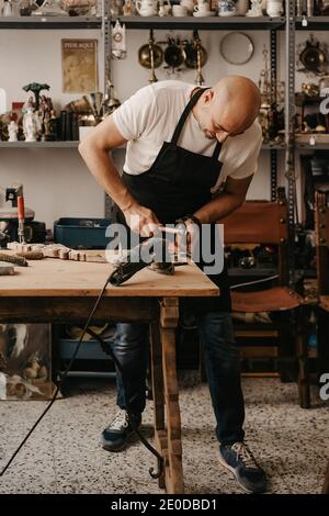 Geschickter reifer Handwerker in Schürze mit Hammer während der Arbeit mit Hölzernes Detail an der Werkbank in der kreativen Werkstatt Stockfoto