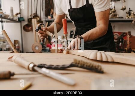 Crop unkenntlich Holzarbeiter in Schürze Schnitzerei Holz mit Meißel und Hammer beim Erzeugen von kunstvollen Details in der Werkstatt Stockfoto