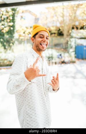 Positive junge bärtige Hipster männlich in gelb Hut und trendy Informelles Hemd lächelnd und Blick auf die Kamera, während Sie dagegen stehen Verschwommenes urbanes Backgro Stockfoto