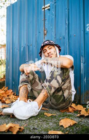 Ganzer Körper von jungen tätowierten männlichen Hipster in informeller Kleidung Sitzt auf dem Boden in der Nähe von Metallzaun und Blick auf die Kamera Stockfoto