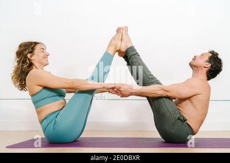 Seitenansicht des lächelnden, flexiblen Paares, das auf der Matte sitzt Navasana und Balancing beim acroyoga im Studio Stockfoto