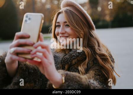 Charmante Weibchen in Baskenmütze und warmen Mantel im Park stehen Und Selfie auf dem Smartphone, während Sie lächeln und das Wochenende genießen Stockfoto