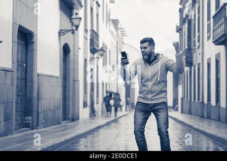 Junger Mann, der mitten auf der Straße auf der verregneten Straße den Sieg feiert. Verrückte Hipster männlichen Modell hält Handy und Faust nach oben. Online-Wette Stockfoto