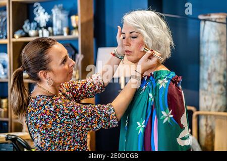 Fokussierte Make-up-Künstler Anwendung Pulver mit Pinsel auf Gesicht Frau mittleren Alters, während sie im Zimmer in der Wohnung steht Stockfoto