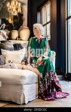 Heitere reife Frau trägt lange Seidenkleid streicheln charmanten Hund Während Sie auf dem Sofa im Luxuszimmer sitzen und wegschauen Stockfoto