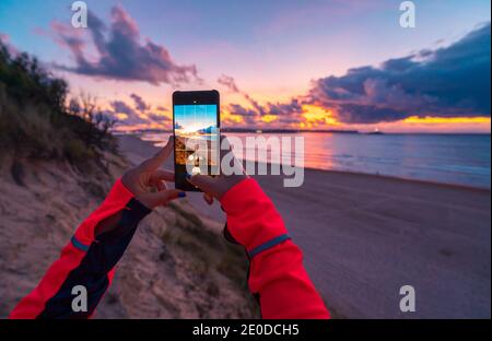 Rückansicht von nicht erkennbaren weiblichen Reisenden mit langen dunklen Haaren In legerer Kleidung fotografieren Sie den atemberaubenden Sonnenuntergang über dem Meer Mit Sandstrand auf smartp Stockfoto