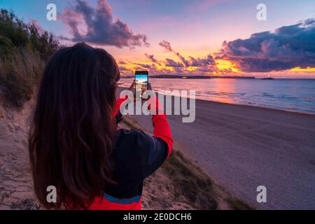 Rückansicht von nicht erkennbaren weiblichen Reisenden mit langen dunklen Haaren In legerer Kleidung fotografieren Sie den atemberaubenden Sonnenuntergang über dem Meer Mit Sandstrand auf smartp Stockfoto