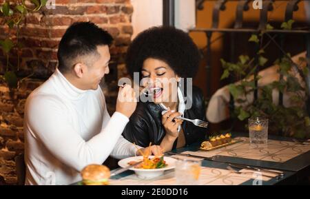 Glücklich asiatische Mann Fütterung fröhlich afrikanisch amerikanische Freundin mit Salat Während des Mittagessens im Café Stockfoto