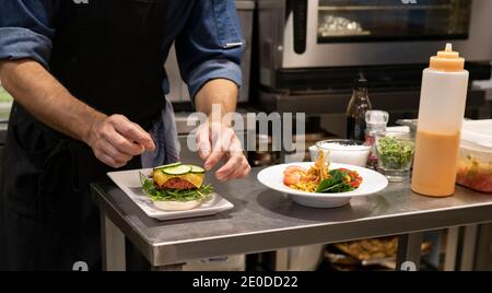 Crop unkenntlich männlichen kochen Zugabe von geschnittenen frischen Gurken Hamburger Mit Fleischschnitzel und Käse während der Zubereitung von Snack im Restaurant Küche Stockfoto