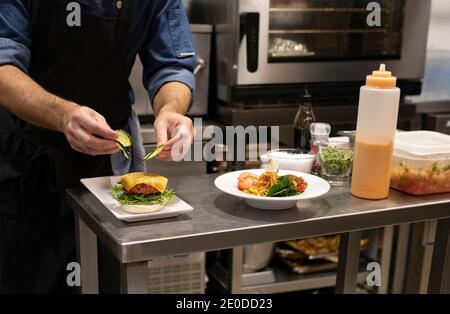 Crop unkenntlich männlichen kochen Zugabe von geschnittenen frischen Gurken Hamburger Mit Fleischschnitzel und Käse während der Zubereitung von Snack im Restaurant Küche Stockfoto