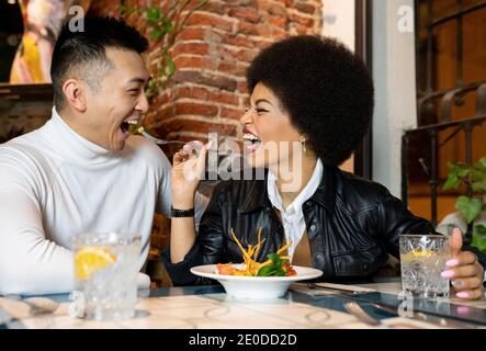 Lachende afroamerikanische junge Frau, die fröhlichen asiatischen Freund mit Salat füttert, während sie sich beim Mittagessen im Café amüsiert Stockfoto