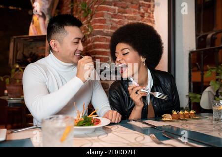 Glücklich asiatische Mann Fütterung fröhlich afrikanisch amerikanische Freundin mit Salat Während des Mittagessens im Café Stockfoto