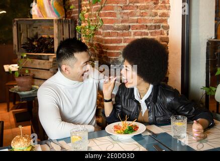 Lachende afroamerikanische junge Frau, die fröhlichen asiatischen Freund mit Salat füttert, während sie sich beim Mittagessen im Café amüsiert Stockfoto