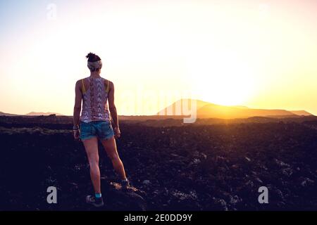 Rückansicht von unkenntlich weibliche Reisende in Casual Outfit stehen Auf unwegsamem felsigen Gelände und bewundern Sie die malerische Landschaft des Sonnenuntergangs Über die Berge Stockfoto