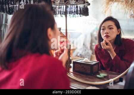 Junge asiatische weibliche Anwendung roten Lippenstift, während im Vordergrund stehen Spiegel im Zimmer mit traditionellem orientalischen Interieur Stockfoto