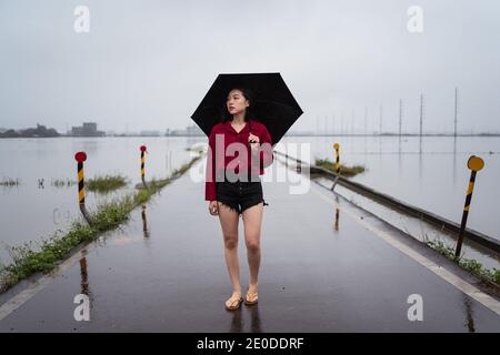 Melancholische Hündin mit Regenschirm, die barfuß auf nasser, leerer Straße läuft An regnerischen Tag unter bewölktem Himmel in Yilan County Stockfoto