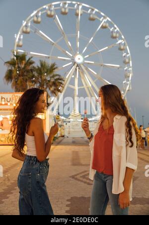 Niedriger Winkel Seitenansicht von glücklich sorglos teen weibliche Freunde Mit Süßigkeiten in den Händen genießen Sommerabend auf Jahrmarkt während Gegen das Riesenrad stehen Stockfoto