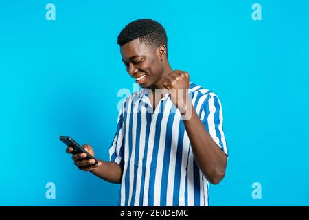 Entzückt afroamerikanischen männlichen Surfen Smartphone und lesen gute Nachrichten Über Business-Projekt beim Stehen mit geballter Faust auf Vivid Blaue Rückseite Stockfoto