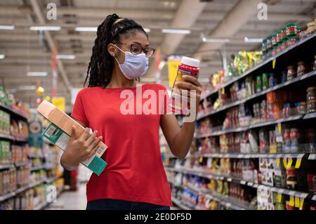 Afro latina junge Frau mit Gesichtsmaske geht Gang und schaut sich Produkte an, während sie im Supermarkt einkaufen Stockfoto