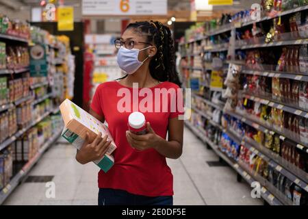 Afro latina junge Frau mit Gesichtsmaske geht Gang und schaut sich Produkte an, während sie im Supermarkt einkaufen Stockfoto