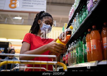 Afro latina junge Frau mit Gesichtsmaske liest das Etikett in der Sodaflasche beim Einkaufen im Supermarkt Stockfoto