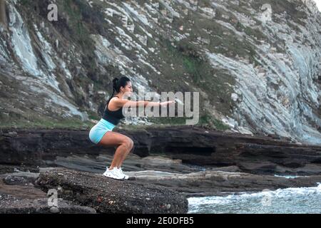 Seitenansicht der fokussierten weiblichen Sportlerin im aktiven Kniebeugen Beim Training an der felsigen Küste nahe am Meer und wegschauen Stockfoto