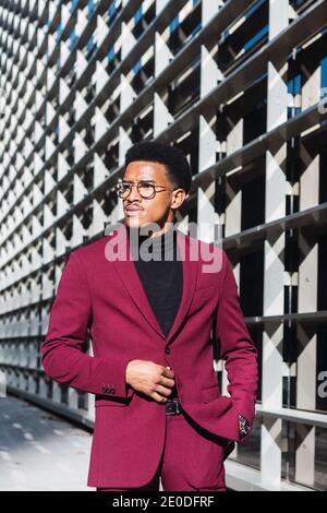 Selbstbewusster junger afroamerikanischer Rüde in modischem Anzug und Brillen Auf der Straße in der Nähe eines modernen Gebäudes mit Glaswänden Sonniger Tag Stockfoto