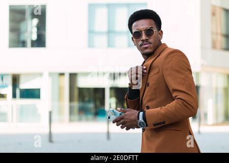 Selbstbewusster afroamerikanischer Mann mit stilvoller Jacke und Sonnenbrille Anpassen Kragen des Hemdes während des Stehens in der Stadt und suchen Bei der Kamera Stockfoto