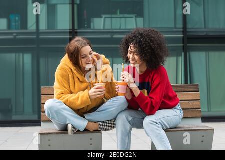 Entzückt multiethnischen Freundinnen in trendigen tragen sitzen auf der Bank In der Stadt und trinken süßen Saft aus Kunststoff zu gehen Tassen beim Betrachen jeder ot Stockfoto