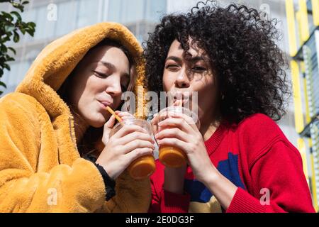 Entzückt multiethnischen Freundinnen in trendigen tragen sitzen auf der Bank In der Stadt und trinken süßen Saft aus Kunststoff zu gehen Tassen beim Betrachen jeder ot Stockfoto