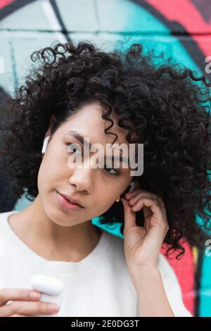 Ruhige afroamerikanische Frau mit Afro Frisur setzen auf wahr Kabellose Kopfhörer und Musikhören auf der Straße der Stadt Stockfoto