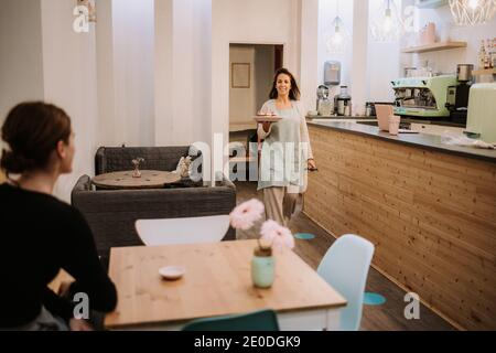Positive weibliche Süßwarenbesitzerin trägt Teller mit Süßigkeiten während des Serviers Dessert für Gäste im gemütlichen Café Stockfoto