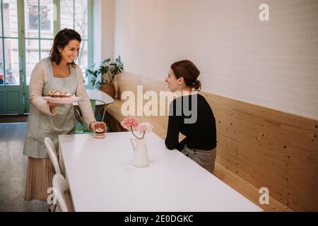 Fröhliche weibliche Konditorin im Gespräch mit lachenden Kunden, während sie lecker serviert Dessert in gemütlicher Cafeteria Stockfoto
