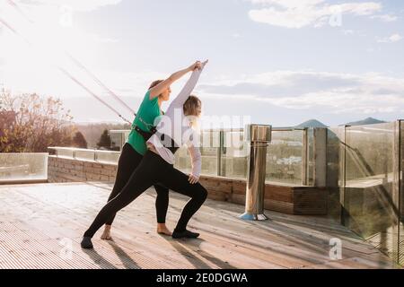 Ruhig Weibchen in Gurten stehen in Virabhadrasana und Stretching Körper Mit Unterstützung von Personal Trainer beim Yoga auf der Terrasse An sonnigen Tag Stockfoto