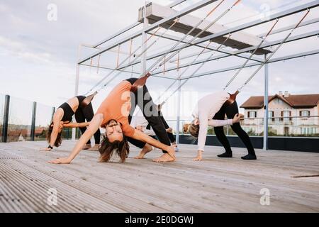 Gruppe von Menschen in Sportbekleidung, die sich auf Riemen lehnen und üben Yoga in Parivrtta Adho Mukha Svanasana auf Holzterrasse Stockfoto