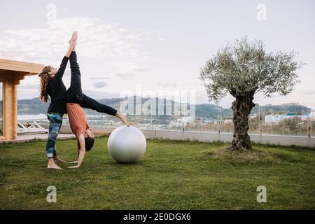 Seitenansicht der weiblichen persönlichen Yoga-Ausbilder helfen Mann mit Handstand Asana lehnt auf fit Ball auf Rasen Stockfoto