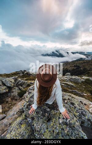 Rückansicht der gesichtslosen weiblichen Touristin im Hut sitzend Stein und Beobachtung landschaftlich Blick auf die Berge an bewölkten Tag Stockfoto