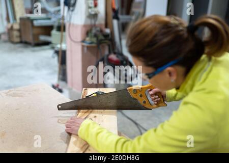 Seitenansicht einer anonymen Zimmermannin Uniform und Brille Scharfe Säge beim Schneiden von Holzdielen in der Werkstatt verwenden Stockfoto