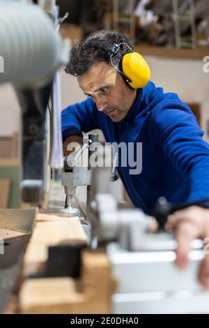 Seitenansicht eines konzentrierten männlichen Zimmermanns mittleren Alters in Kopfhörern Arbeiten an der Tischsägemaschine in Schreinerei Stockfoto