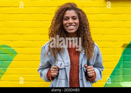 Optimistische junge schwarze weibliche Millennial mit lockigem Haar in stilvoll Die Kleidung lächelt, während man die Kamera an die gelbe Wand anschaut Stockfoto