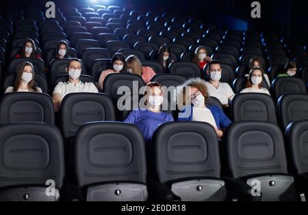 Frontansicht von Menschen, die während der Weltpandemie Film genießen, Blick auf die große Leinwand. Junge Zuschauer sehen Film im Kino, tragen weiße Gesichtsmasken. Soziale Distanzierung, Pandemiekonzept. Stockfoto